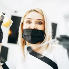 Uśmiechnięta dentystka w masce przygotowuje się do lakierowania zębów, trzymając lampę i narzędzia.