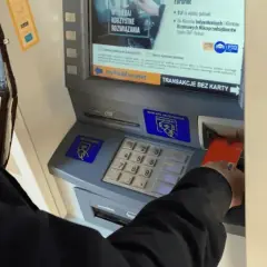 Osoba wkłada kartę do bankomatu, by wypłacić pieniądze. Widoczny portfel z kartami.