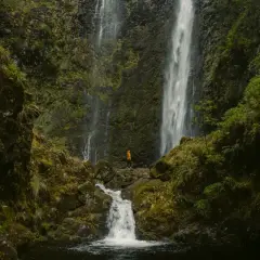 A lone hiker in an orange jacket stands at the base of the majestic Caldeirão Verde waterfall, a breathtaking sight on this lush hike.