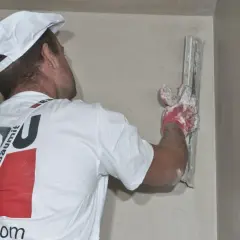 Murarz w białej czapce i koszulce z logo Baumit nakłada tynk na ścianę za pomocą kielni.