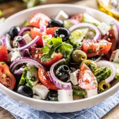 Ensalada griega fresca con tomates, pepinos, cebolla morada, aceitunas negras y queso feta, servida en un bol blanco.