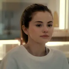 Selena Gomez w białej bluzie, z włosami związanymi w kucyk, patrzy w bok zamyślona.