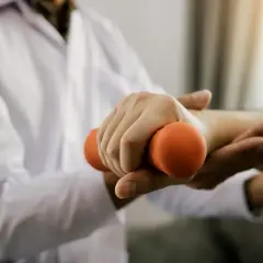 Fizjoterapeuta pomaga pacjentowi z brakiem siły w rękach, ćwicząc z małym, pomarańczowym ciężarkiem.