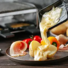 Formaggio fuso in padella, filante e dorato, viene versato su patate e salumi. Un piatto invitante e gustoso.