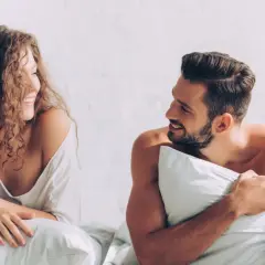Coppia sorridente a letto, con la donna dai capelli ricci che guarda l'uomo con barba, un momento intimo prima di fare sesso.