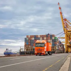 Ein orangefarbener LKW fährt an einem Containerschiff und einem Hafenkranausleger vorbei. Das zeigt, was eine Spedition leistet: Güter transportieren.
