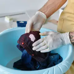 Hände mit Handschuhen behandeln ein Kleidungsstück mit einem rosa Fleck in einer blauen Schüssel, um **Flecken entfernen** zu können.