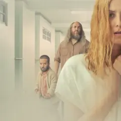 Kobieta z blond włosami i grupa ludzi w korytarzu. Wszyscy wyglądają na zamyślonych, jakby podążali za krzywymi liniami boga.