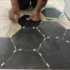 Hands place dark hexagonal tiles over existing floor tiles, using white spacers to ensure even gaps.