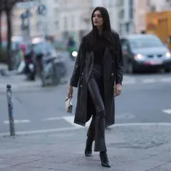 Kobieta w czarnym płaszczu i skórzanych spodniach idzie ulicą.