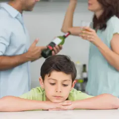 Smutny chłopiec opiera głowę na rękach, podczas gdy rodzice w tle piją alkohol. Obraz symbolizuje trudne dzieciństwo i problemy związane z dda.