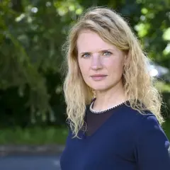 Wiktoria Grodecka z falowanymi blond włosami i niebieskimi oczami, w granatowym swetrze, na tle zieleni.