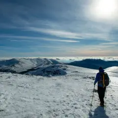 Senderismo en la nieve de Peñalara. Un excursionista con mochila y bastones contempla un paisaje montañoso cubierto de nieve bajo un cielo azul.