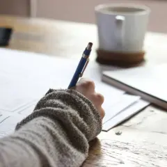 Ręka pisze w dzienniczku uczuć, obok kubek kawy i notatnik.