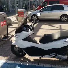 Na parkingu stoi biały skuter. Obok znajduje się automat z oznaczeniami "P" i symbolem motocykla, co sugeruje, że motocykle płacą za parking.