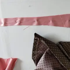 Różowa taśma z agrafkami, przygotowana do poszerzenia sukienki po bokach. Obok leży wzorzysta tkanina.