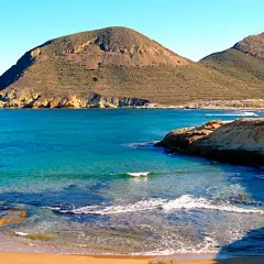 Aguas turquesas bañan la costa rocosa de el playazo las negras, con montañas áridas de fondo bajo un cielo azul.
