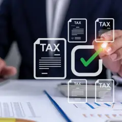 Dłoń zaznacza zielonym znacznikiem dokumenty z napisem "TAX" na tle wykresów i laptopa.