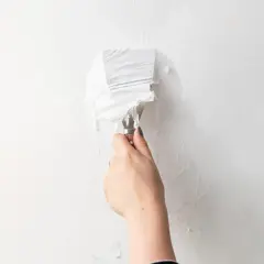 Mano con spatola che rimuove strati di carta da parati bianca. Un passo fondamentale per capire come togliere la carta da parati.