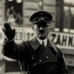 Adolf Hitler winkt. Er trug eine Uniform und einen Hut. Hinter ihm ist eine Tafel mit der Aufschrift "Zahnatelie". Was hat Hitler gemacht? Er hat den Zweiten Weltkrieg begonnen.
