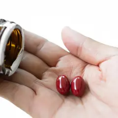 Zwei rote Kapseln liegen in einer Hand, während eine braune Flasche entleert wird. Fragen zur Sicherheit von Astaxanthin krebserregend bleiben offen.