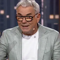 Mężczyzna w okularach i jasnej marynarce gestykuluje rękami na tle miejskiego krajobrazu.