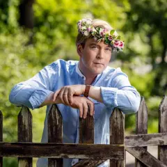 En man med blomsterkrans på huvudet lutar sig mot ett staket. En sommarprat recension som fångar essensen av svensk sommar.