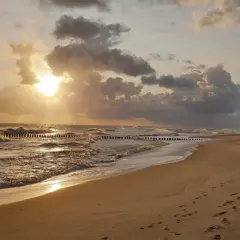 Piękny zachód słońca we Władysławowie. Słońce przebija się przez chmury, tworząc złote refleksy na falach Bałtyku. Na piaszczystej plaży widać ślady stóp.