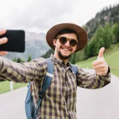 Uśmiechnięty turysta w kapeluszu robi sobie selfie z kciukiem w górę na tle gór. Widać, jak z łatwością zrobić selfie.