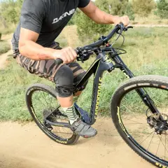 A rider on a black and yellow single fork mountain bike navigates a dusty trail, wearing knee pads and camouflage shorts.