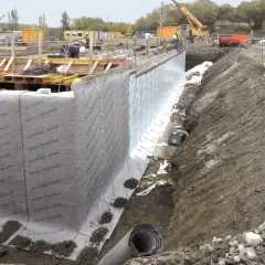 Baustelle mit frisch verlegter Noppenbahn wie rum. Betonfundament und Baugrube sind sichtbar.