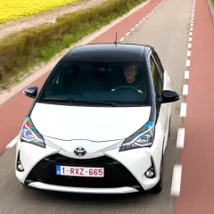 Biały Toyota Yaris Sedan z czarnym dachem jedzie drogą wśród pól żółtych kwiatów i czerwonych tulipanów.