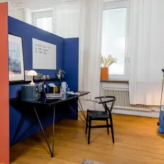 Ein blauer Raumteiler trennt den Arbeitsplatz im Wohnzimmer ab. Ein Schreibtisch mit Laptop und Lampe steht bereit.