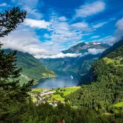Widok na fiord podczas rejsu do Norwegii. Statek wycieczkowy płynie po spokojnej wodzie, otoczonej zielonymi górami i chmurami.