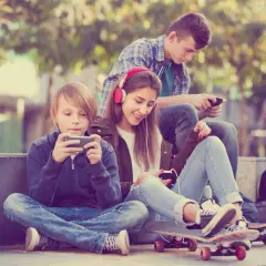 Trójka nastolatków pochłonięta smartfonami, obok deskorolki. Czy to początek uzależnień behawioralnych?