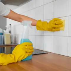 Ręce w żółtych rękawiczkach czyszczą białe płytki w łazience. Jedna ręka trzyma butelkę z niebieskim płynem, druga gąbkę. To pokazuje, czym myc płytki w łazience.