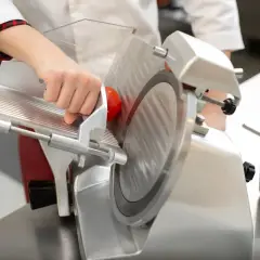 Mano che spinge un pomodoro su un'affettatrice. Per capire come pulire affettatrice, guarda attentamente la lama e il carrello.