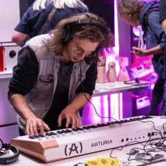 Młody mężczyzna w słuchawkach gra na keyboardzie Arturia. W tle widać sprzęt audio i innych ludzi, tworząc atmosferę pracy w wytwórni muzycznej.