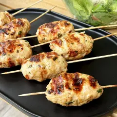 Szaszłyki z mięsa mielonego, apetycznie przypieczone na grillu, leżą na czarnym talerzu. Obok widać miskę z sałatką.