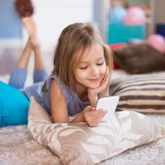 Uśmiechnięta dziewczynka leży na podłodze, przeglądając telefon. Czy rodzice mogą czytać wiadomości swojego dziecka?