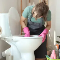 Kobieta w różowych rękawiczkach czyści toaletę, bo w łazience smierdzi. Obok stoją środki czystości i rolki papieru toaletowego.