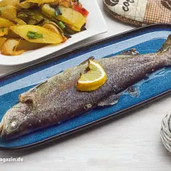 Eine ganze Forelle im Backofen, garniert mit Zitrone, auf einem blauen Teller. Daneben eine Portion Ofenkartoffeln und Paprika.