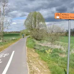 Rowerzysta jedzie ścieżką rowerową przez zielone pola. Pomarańczowy znak wskazuje kierunek do Kotbacza i Szczecin-Śmirdnica.