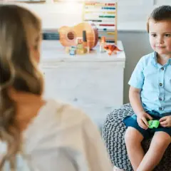 Chłopiec w niebieskiej koszuli siedzi na pufie, rozmawiając z psychoterapeutką.