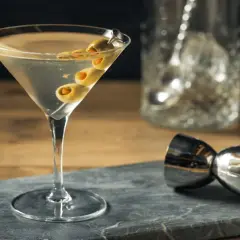Un verre de martini gin, garni de trois olives, prêt à être dégusté. Un doseur à cocktail et un pichet à glaçons sont visibles en arrière-plan.