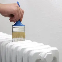 Dłoń z pędzlem maluje biały grzejnik aluminiowy. Odświeżanie wnętrza.