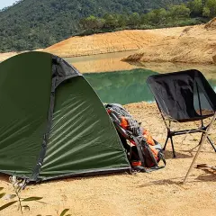 Tienda de campaña verde junto a un lago, con una silla plegable negra y mochilas de transporte.