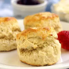 Tre gyllene engelska scones på en tallrik med jordgubbar och grädde i bakgrunden. Perfekt för en mysig fika.