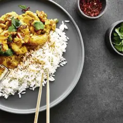 Ein Teller mit Reis und einem köstlichen Thai Curry, garniert mit frischem Koriander. Daneben kleine Schälchen mit Chiliflocken und Koriander.