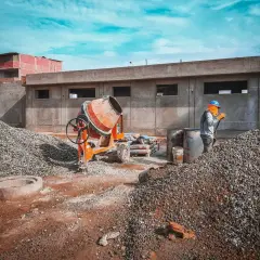 Budowa w toku: robotnik z łopatą przy stercie kruszywa, betoniarka i worki z cementem. Przygotowanie podsypki cementowo piaskowej.